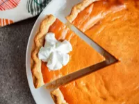 A pumpkin pie on a plate with a slice cut and topped with whipped cream.