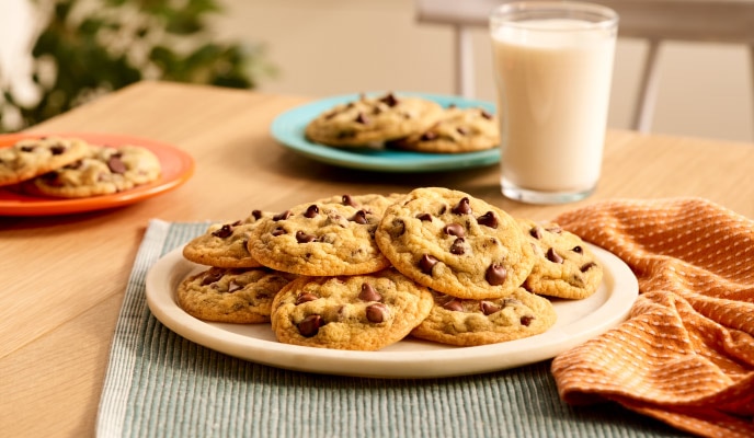 Mesa de madera con un plato grande con una pila de galletas con chispas de chocolate, un vaso de leche y más galletas.